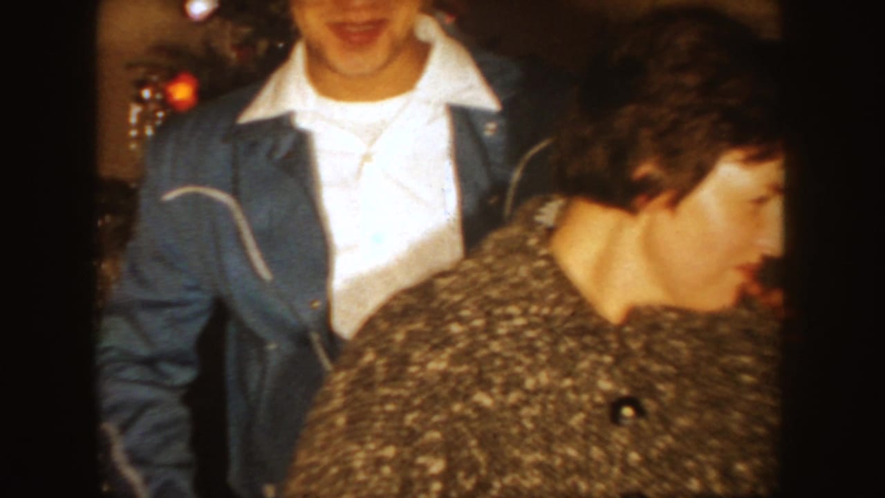 Young man in a blue jacket stands near a woman in a patterned coat by a Christmas tree.