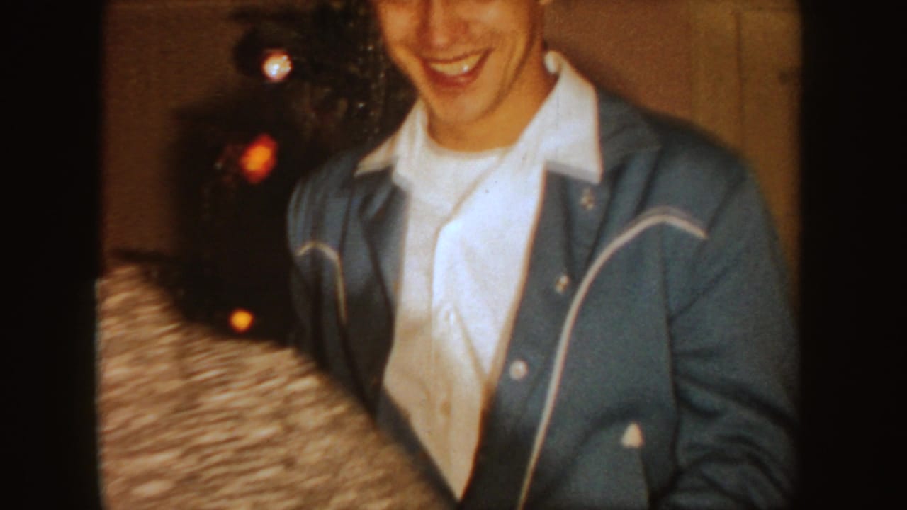 Smiling young man in a blue jacket stands beside a Christmas tree indoors.
