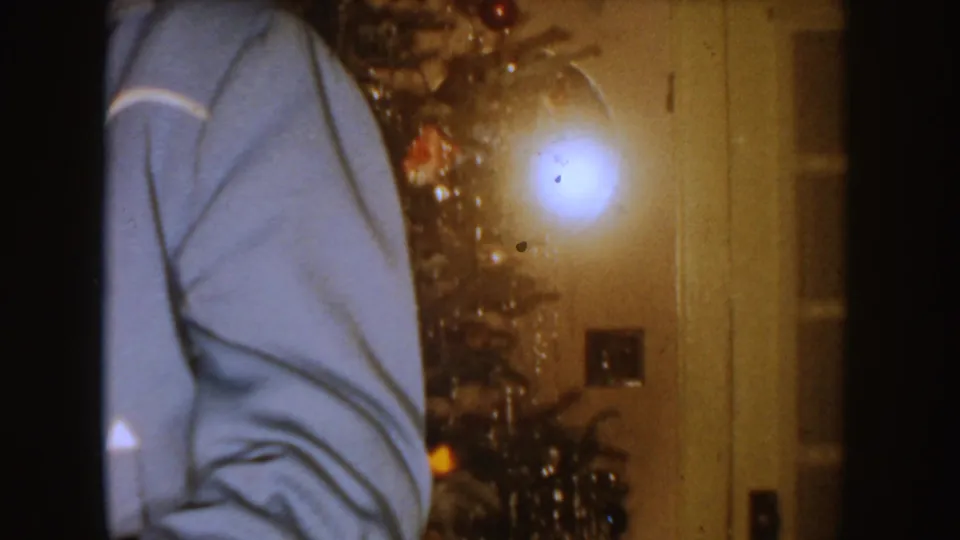 The camera catches a partial figure in a blue jacket, a glowing Christmas tree, and a doorway inside the South Dakota home.