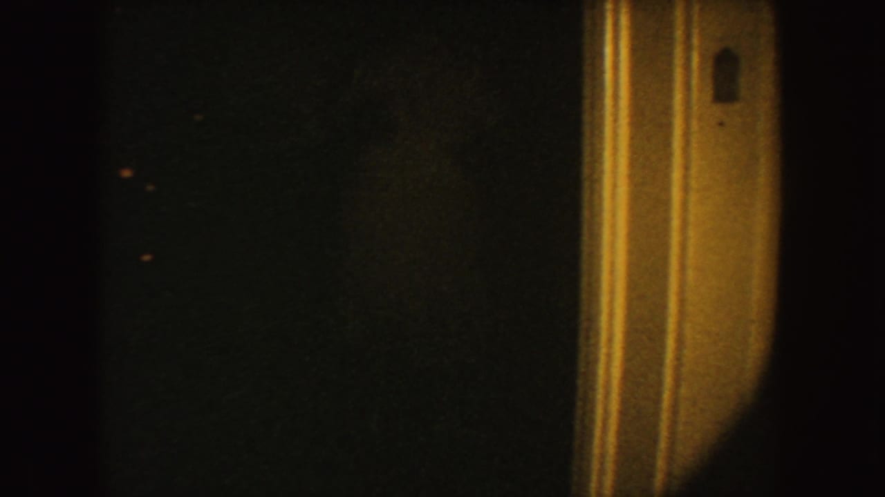 1954 South Dakota 8mm frame showing a tan doorway jamb with a small gray switch plate in dim warm light.