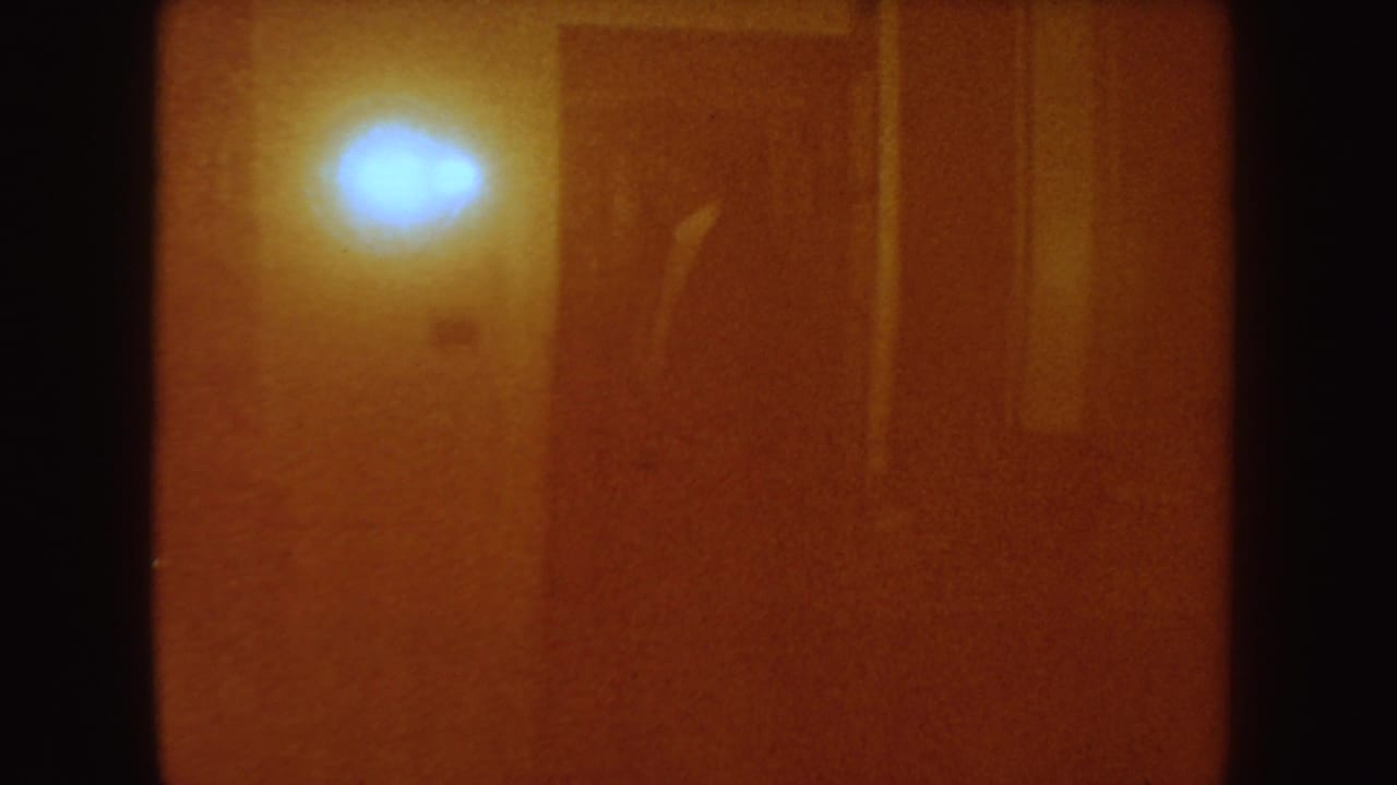 A warm, dimly lit interior from the South Dakota home movie, with an amber wall, bright sconce, dark doorway edge, and heavy film grain.
