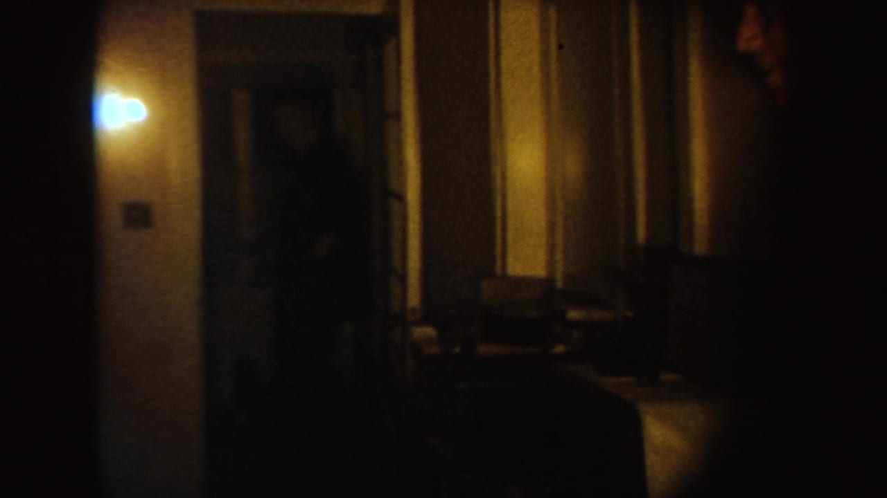 Two adult men in suits appear in a dim South Dakota interior hallway, partly obscured by low light, doorway edges, and grainy 8mm exposure.
