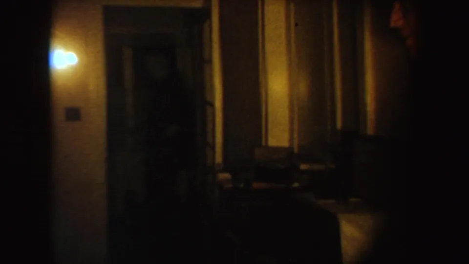 Two adult men in suits appear in a dim South Dakota interior hallway, partly obscured by low light, doorway edges, and grainy 8mm exposure.