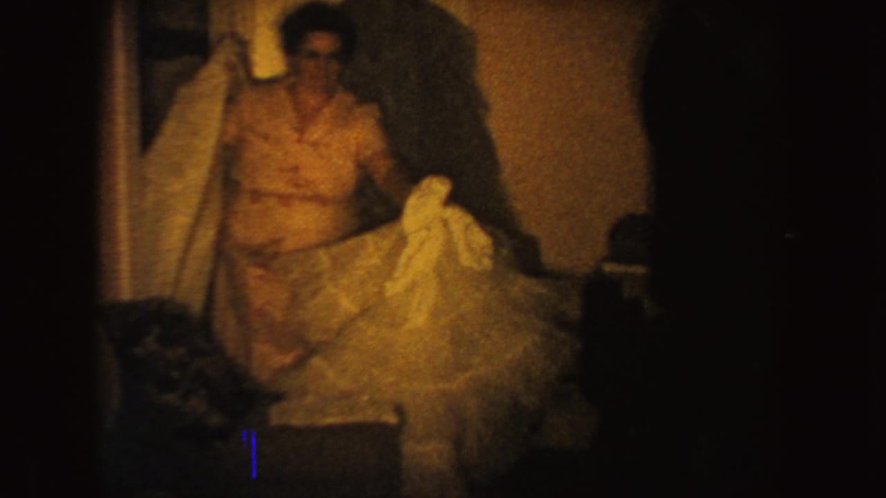 In 1954 South Dakota home-movie footage, a woman in a patterned dress holds a voluminous tulle gown inside a dim private room.