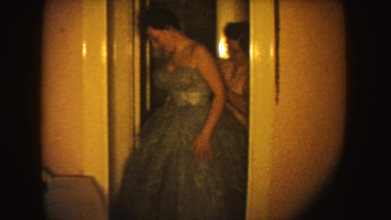 A younger female in a blue-green tulle gown stands at a doorway as an older woman assists, recorded as 1954 South Dakota home-movie footage.