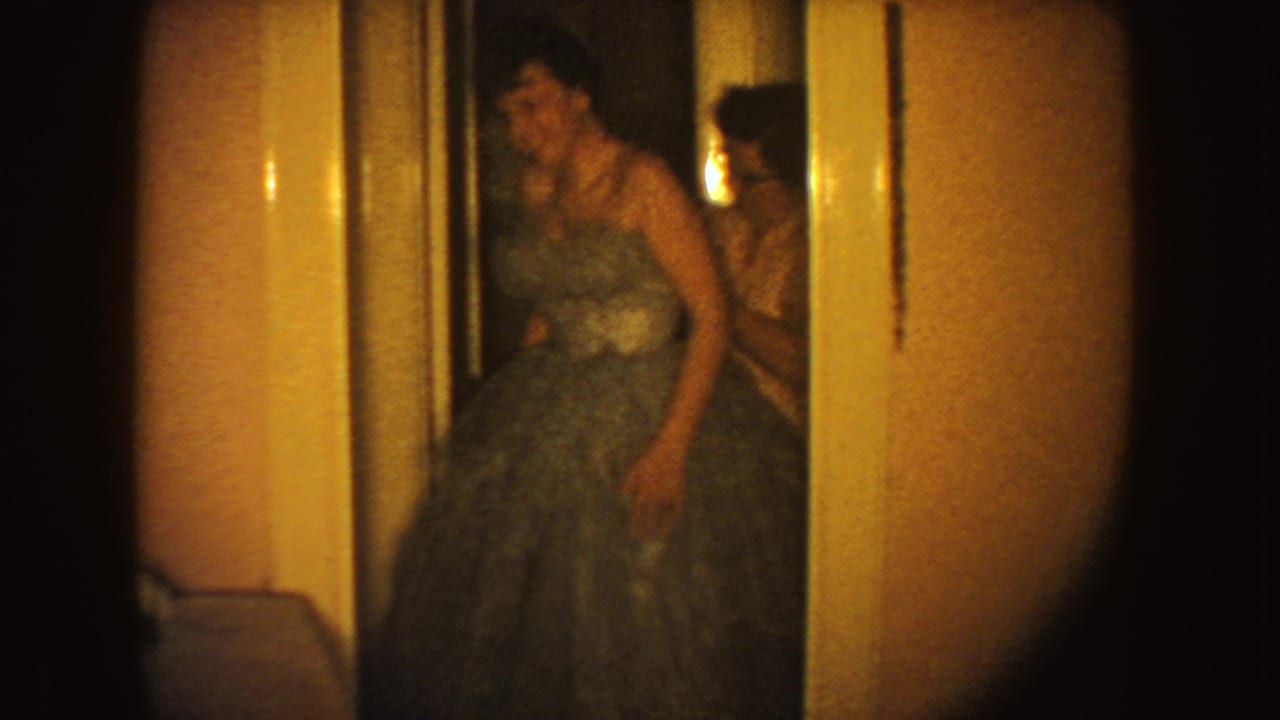 The younger female pauses in the voluminous gown near a doorway, with an assisting woman partly visible behind her in the private interior.