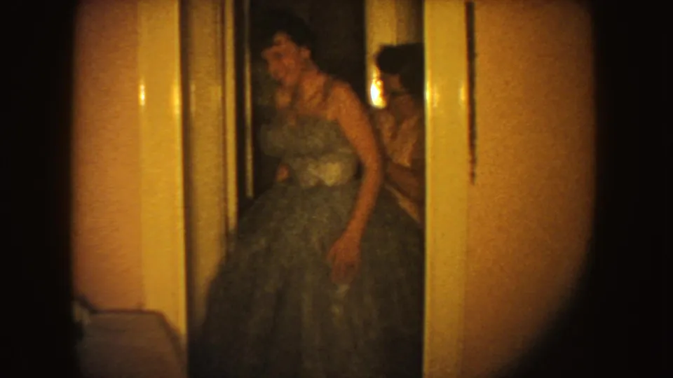 The younger female pauses in the voluminous gown near a doorway, with an assisting woman partly visible behind her in the private interior.