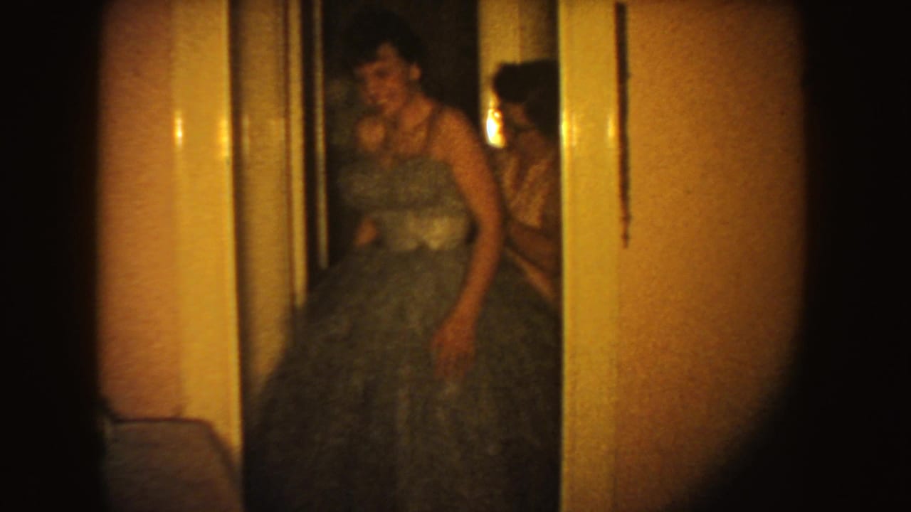 In a 1954 South Dakota home-movie frame, a woman in a voluminous blue gown appears to adjust her dress in a warm hallway.