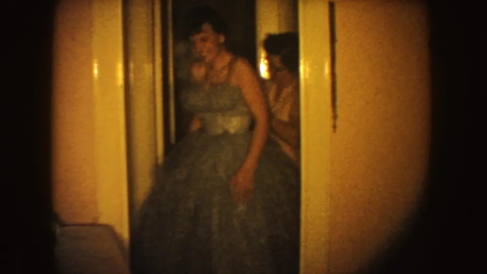 In a 1954 South Dakota home-movie frame, a woman in a voluminous blue gown appears to adjust her dress in a warm hallway.