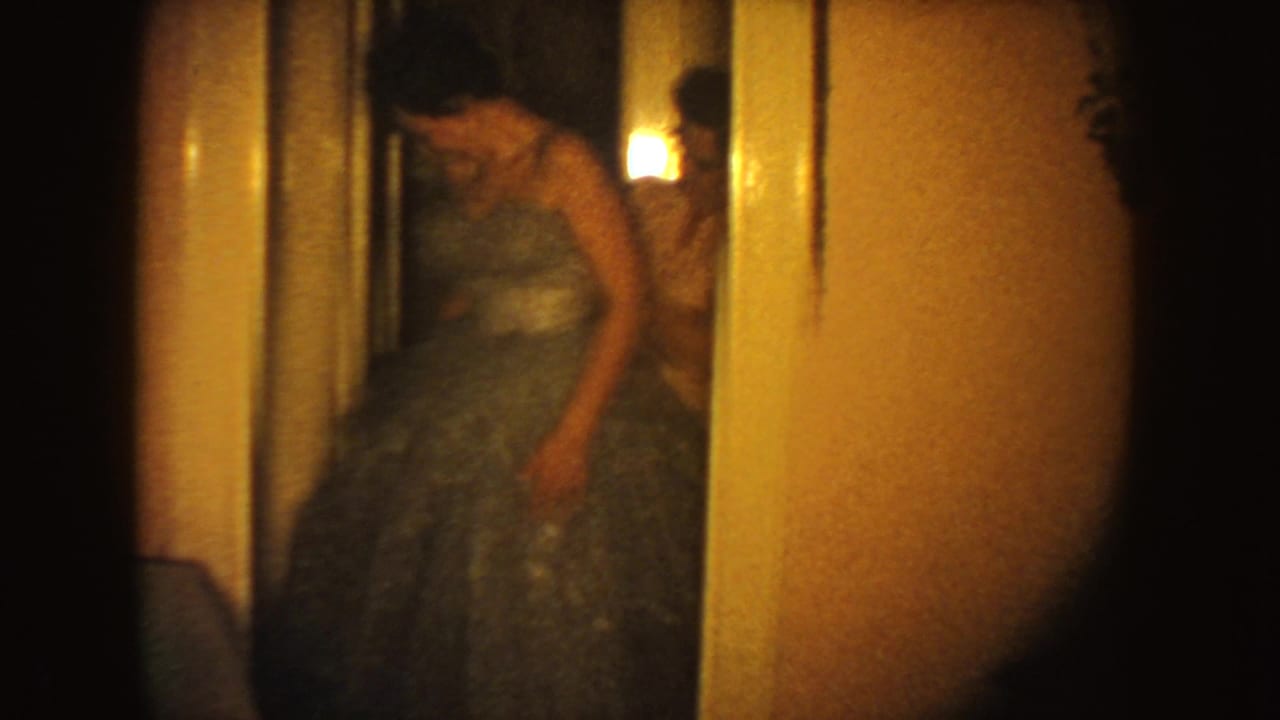 The woman in the blue formal gown looks down as someone behind her appears to adjust the dress in a warmly lit interior.