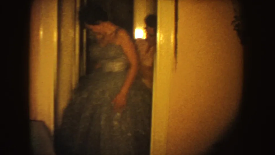 The woman in the blue formal gown looks down as someone behind her appears to adjust the dress in a warmly lit interior.