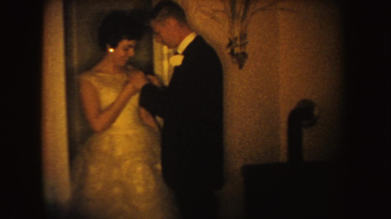 A woman in a light formal gown stands beside a tuxedoed man, likely during wedding preparations in the supplied 1954 South Dakota footage.