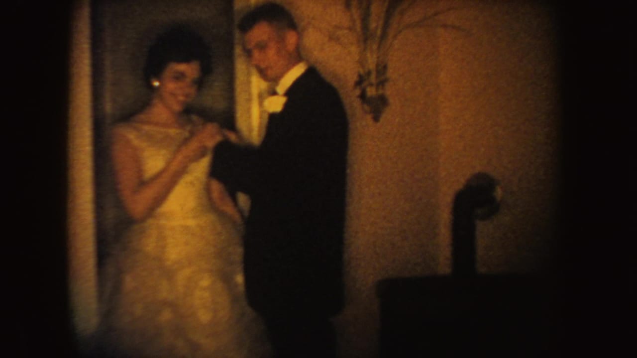 A woman in a light formal gown smiles beside a tuxedoed man in the warm, grainy 1954 South Dakota home-movie scene.