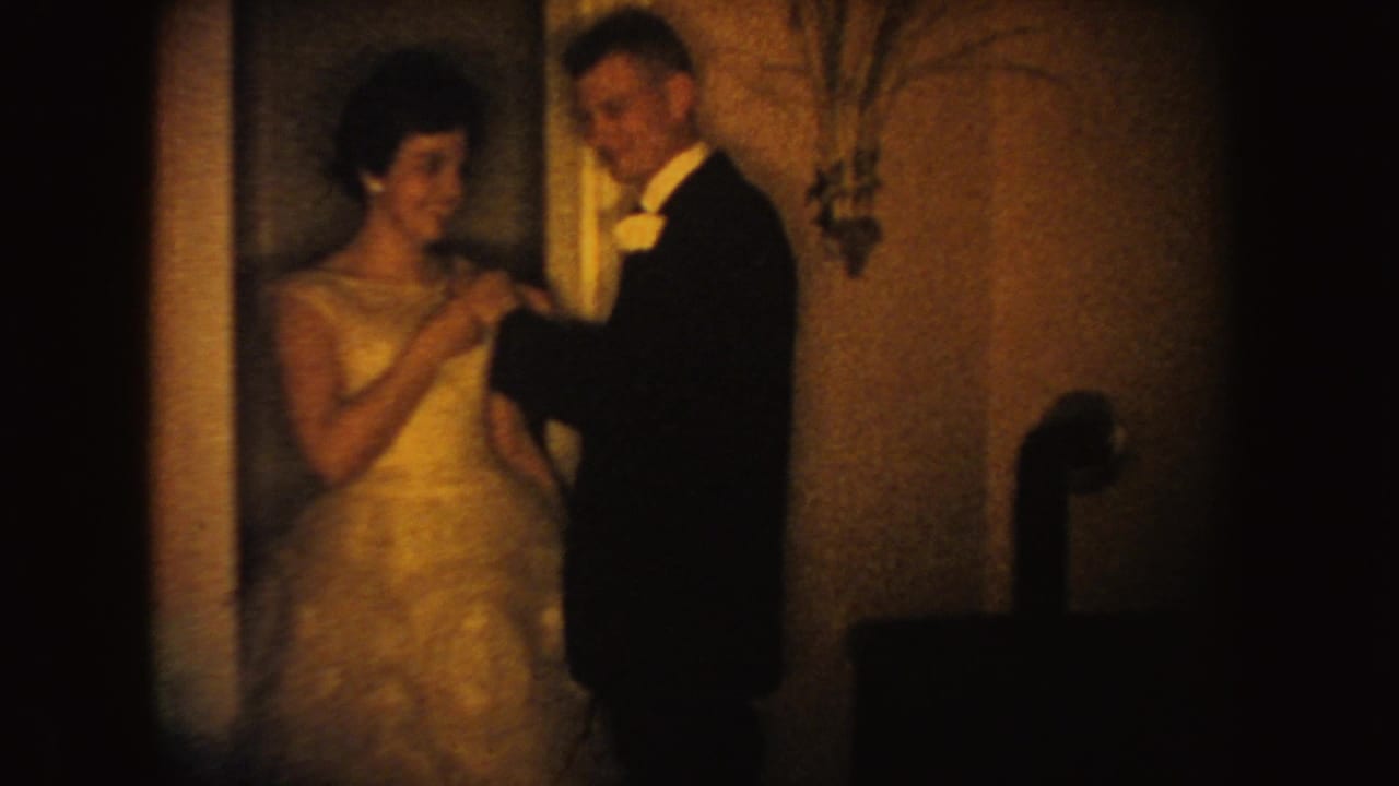 In 1954 South Dakota 8mm footage, a groom in dark formalwear pins a flower to a bride's light wedding dress indoors.