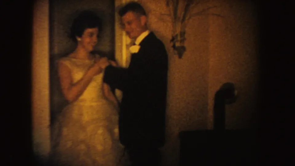 In 1954 South Dakota 8mm footage, a groom in dark formalwear pins a flower to a bride's light wedding dress indoors.