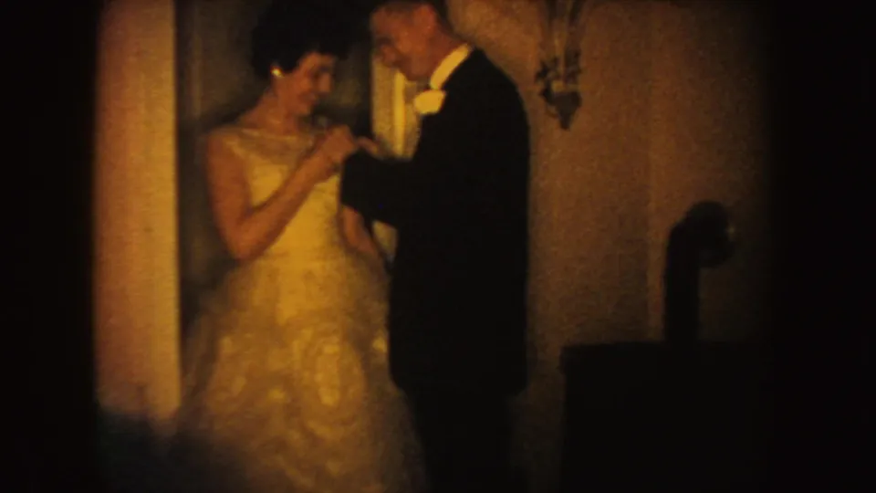 The bride smiles as the groom works near her shoulder, adjusting a flower on her dress in warm 1950s home-movie light.