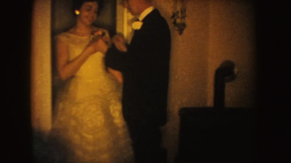 The bride looks upward while the groom finishes with the flower on her dress, filmed indoors on warm, grainy 8mm stock.