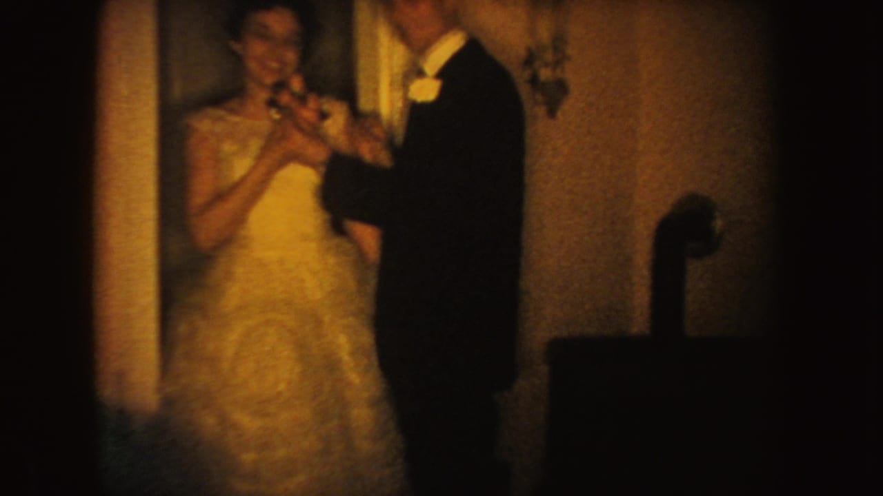 A bride in a light gown and groom in dark formalwear dance inside a South Dakota home-movie wedding scene from 1954.