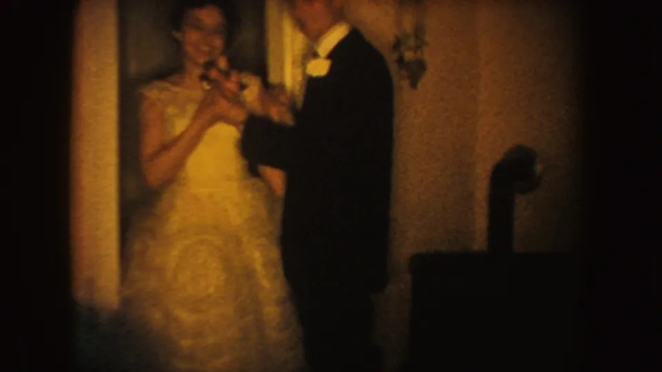 A bride in a light gown and groom in dark formalwear dance inside a South Dakota home-movie wedding scene from 1954.