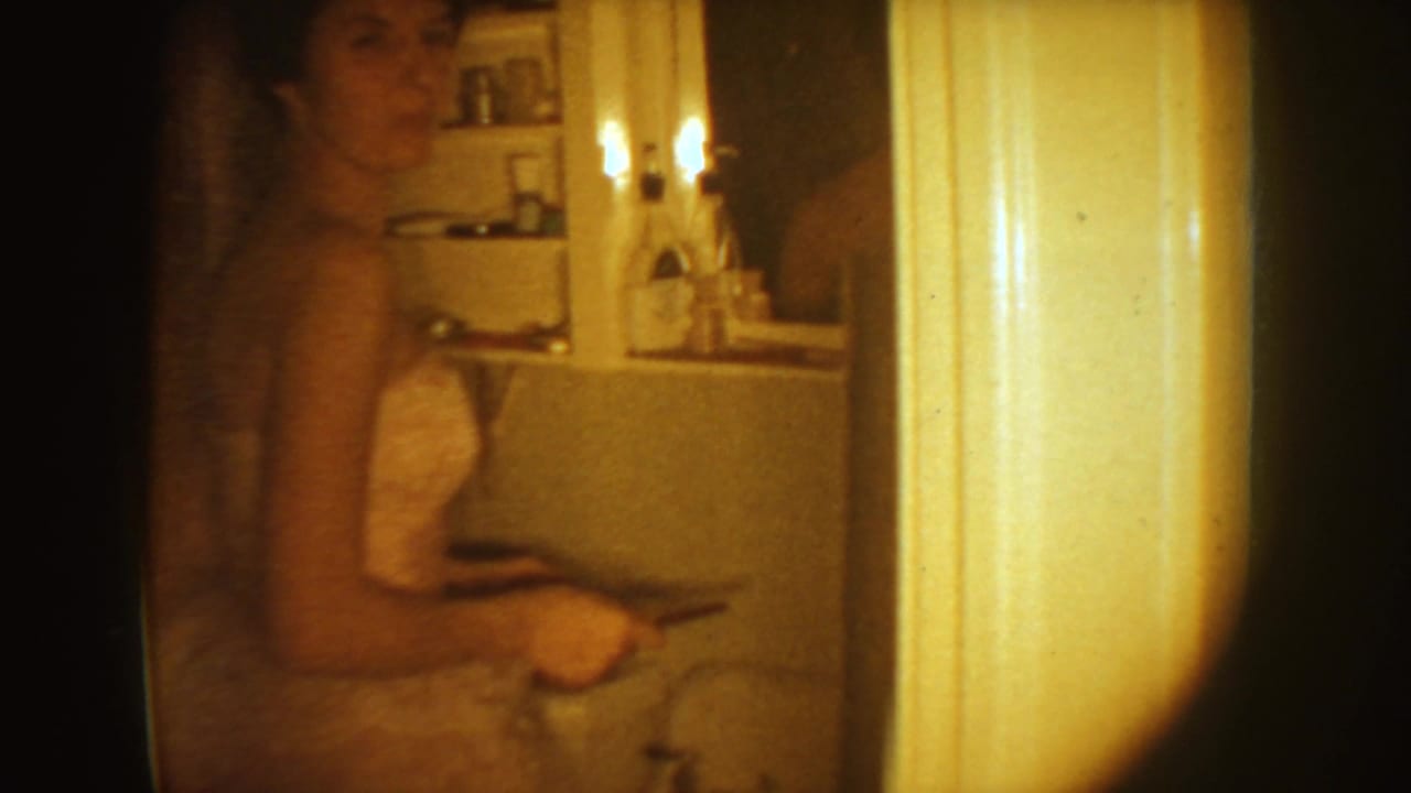 The bride stands in a bathroom interior, holding a small object near a sink and medicine cabinet in the 1954 South Dakota home movie.