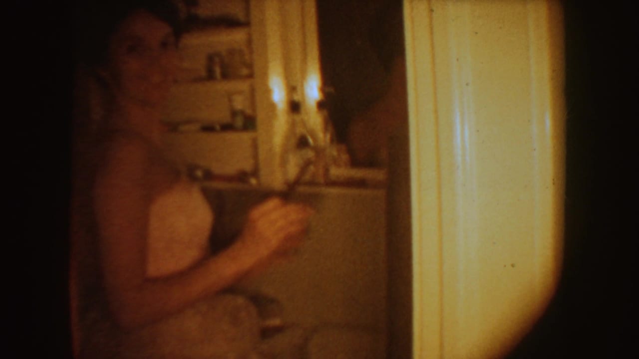 Woman in a white strapless gown standing in a dim bathroom near a mirror or cabinet.
