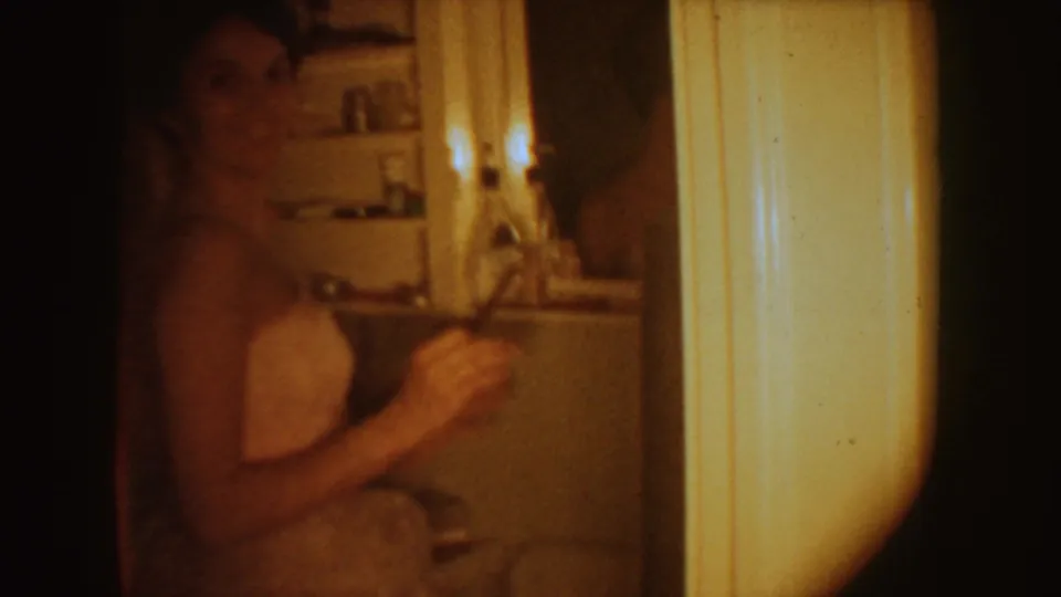 1954 South Dakota 8mm frame of a woman in a white strapless gown in a dim private bathroom, with shelves and pale trim visible.