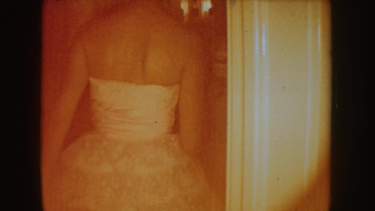 White gown seen through a doorway or mirror area in a warm orange-tinted interior.