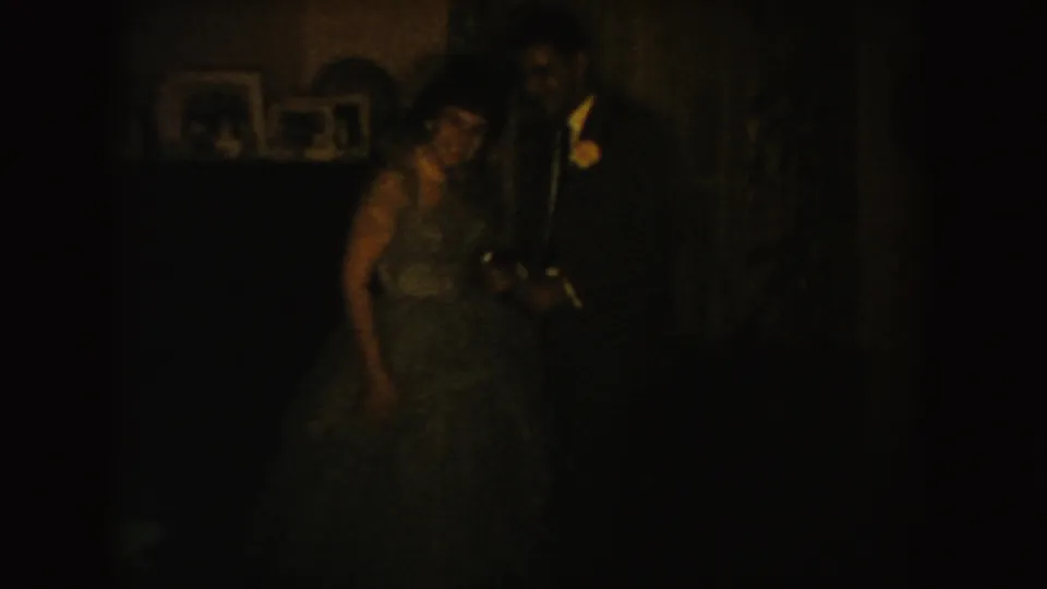 In a dim South Dakota home-movie frame, a woman in a long dress stands close to a man in a dark suit near framed photos.