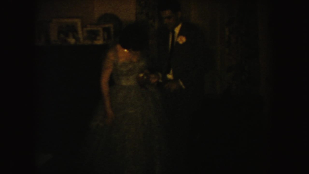Dim frame with bride and groom standing indoors near framed pictures.