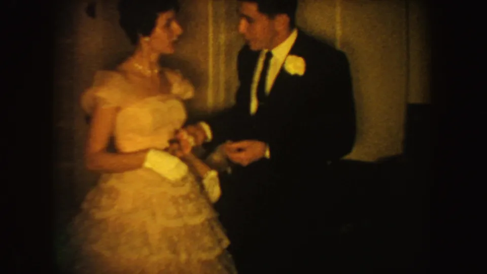 1954 South Dakota home-movie frame showing a bride in a ruffled gown and groom in a dark suit holding hands indoors.