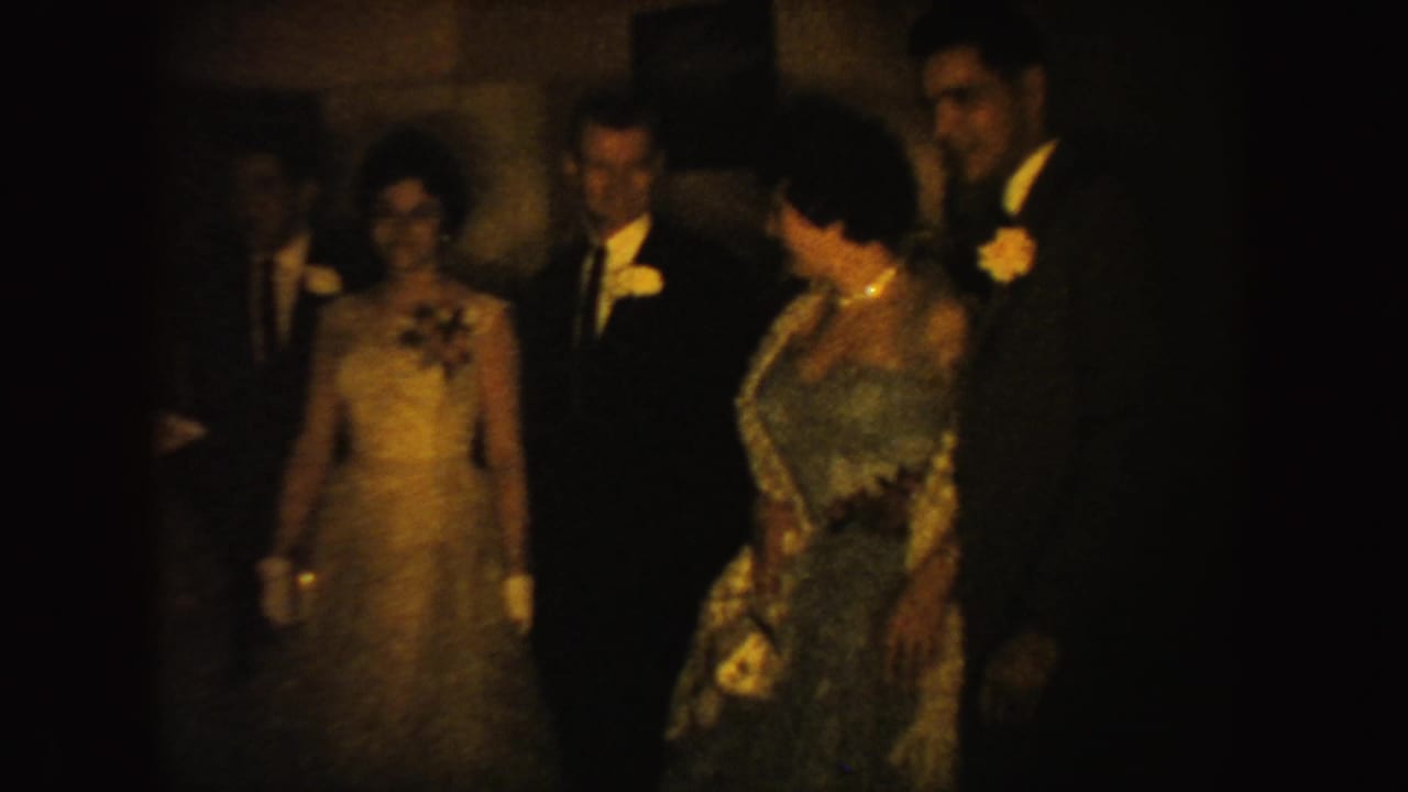 Formal guests in gowns and dark suits stand together in dim indoor light.