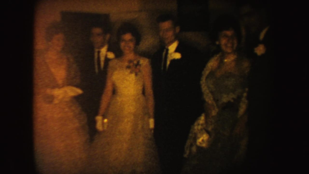 Formal adults in 1954 South Dakota stand in a line, with gowns, suits, gloves, and corsages visible in warm indoor light.