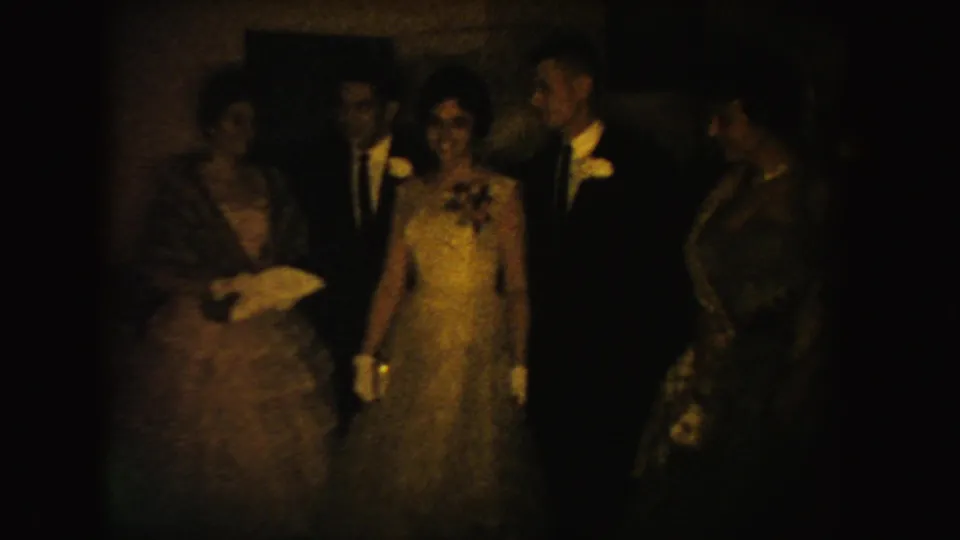 South Dakota 1954 home-movie frame with formal guests clustered indoors, including gowns, suits, gloves, flowers, and soft amber exposure.