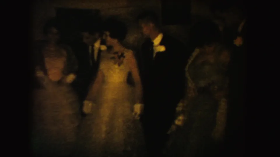 A 1954 South Dakota formal-event frame shows adults in evening clothes shifting position in grainy, warm 8mm footage.