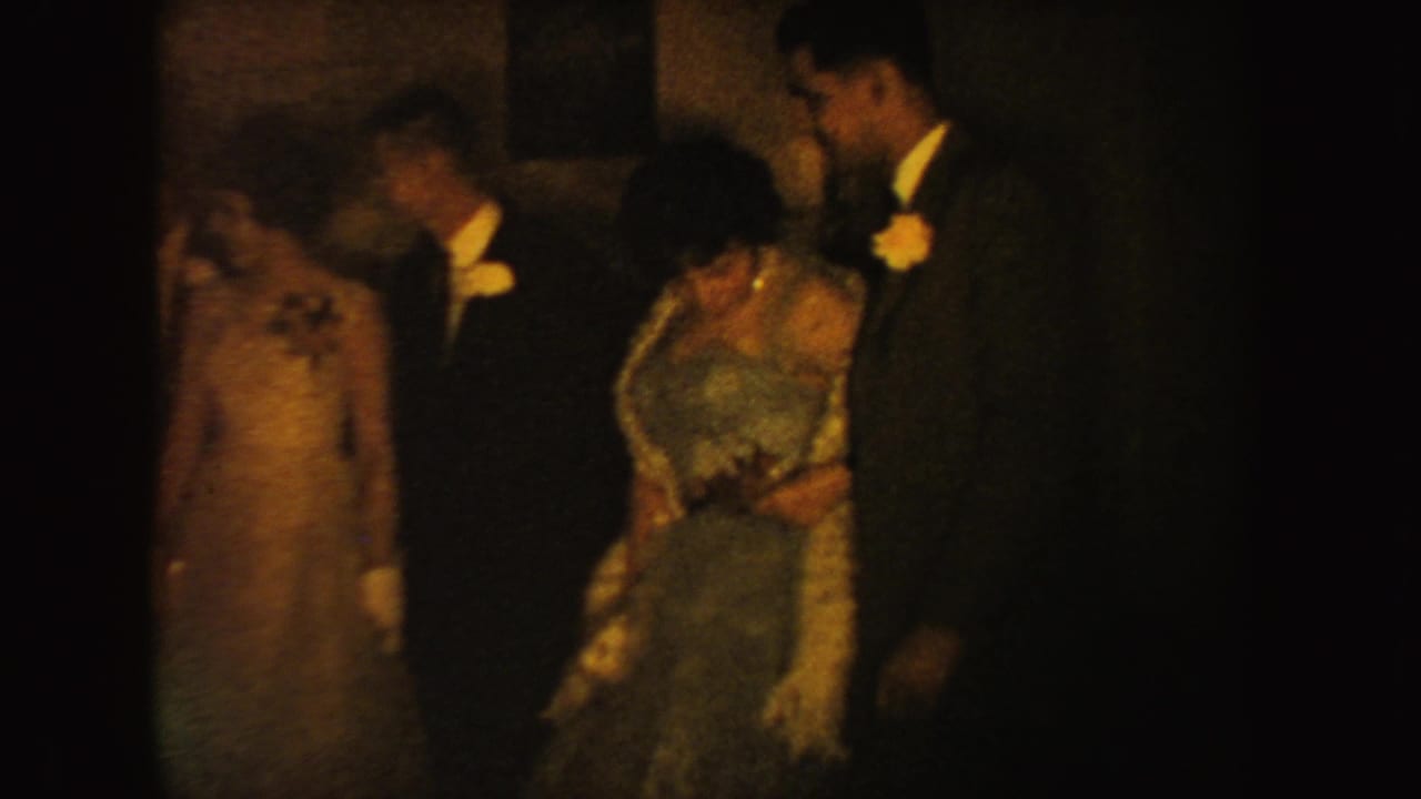 In 1954 South Dakota home-movie footage, two formal couples stand near a dark doorway, with dresses, white gloves, suits, and boutonnieres visible.