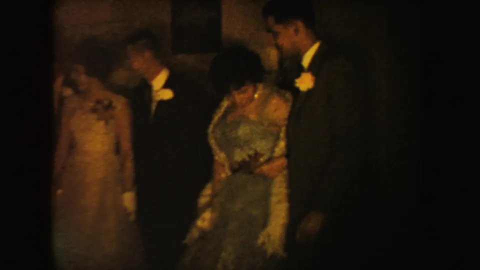 In 1954 South Dakota home-movie footage, two formal couples stand near a dark doorway, with dresses, white gloves, suits, and boutonnieres visible.