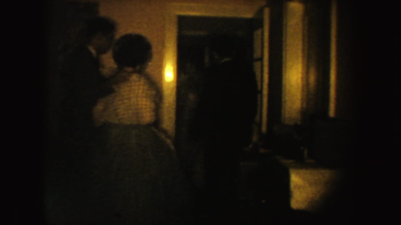 A man and woman stand close together in a dark room near an open doorway.