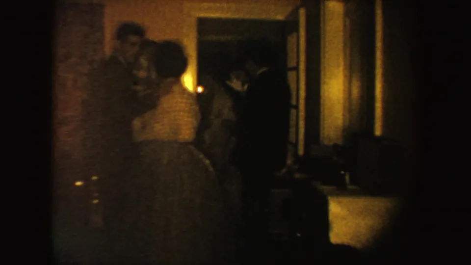 A woman in a full formal dress approaches others near a doorway, with a wall sconce lighting the dim 1954 South Dakota interior.