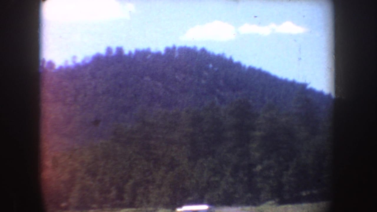 1954 South Dakota home-movie frame showing a wooded hillside, bright sky, and a distant parked automobile near the lower edge.