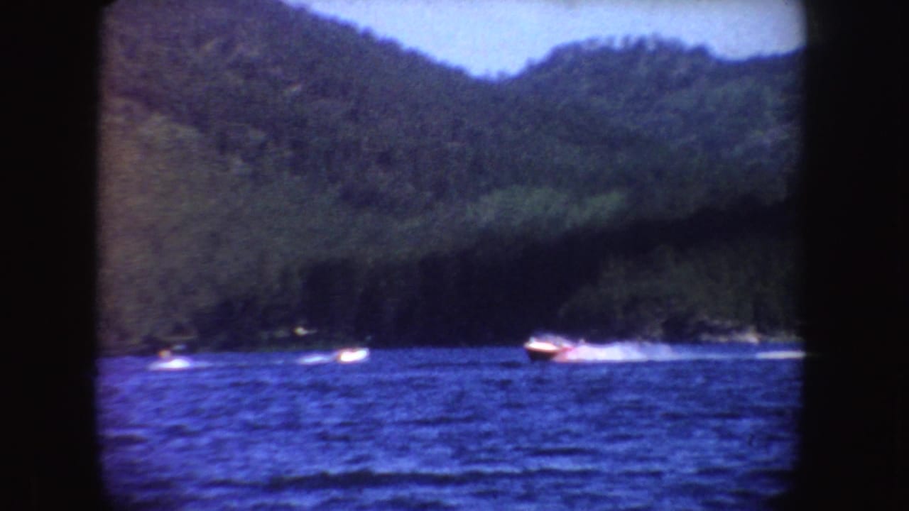 Small speedboats move across a blue lake below wooded hills.