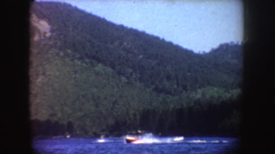 Midcentury 8mm frame of a speedboat on a South Dakota lake, with choppy blue water and a forested slope behind it.