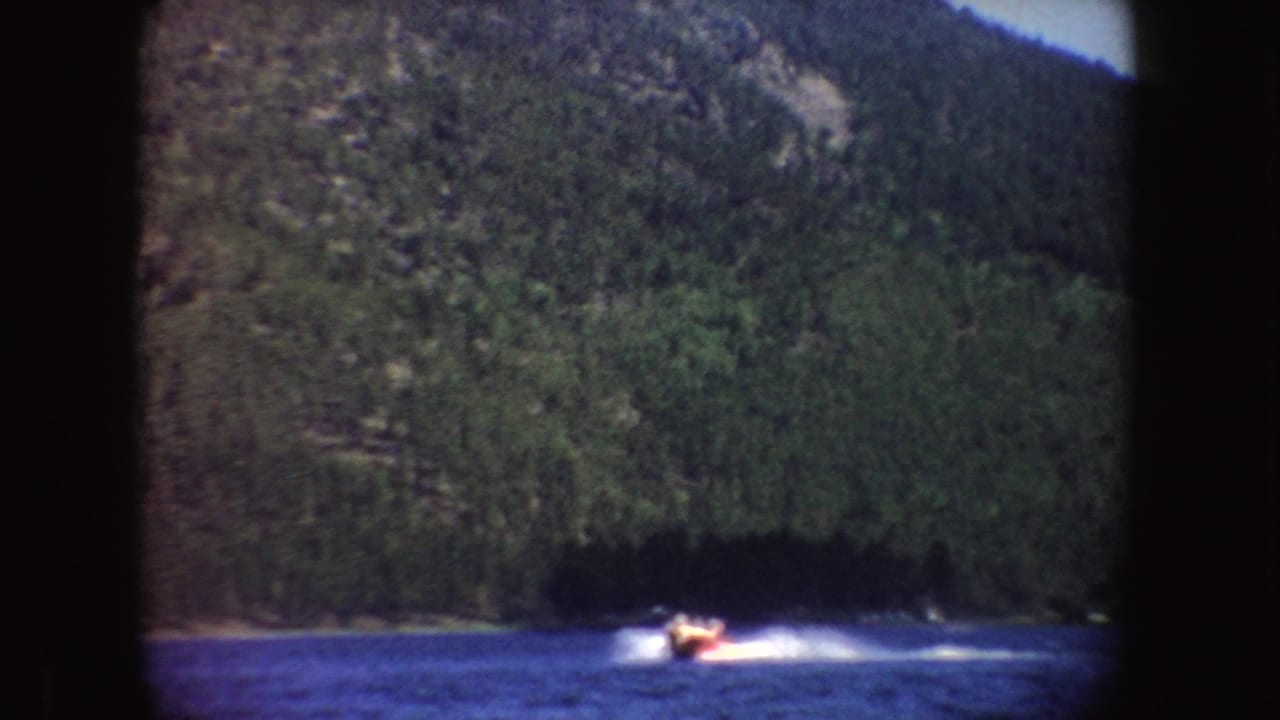 Speedboat crossing a blue lake below forested hills.