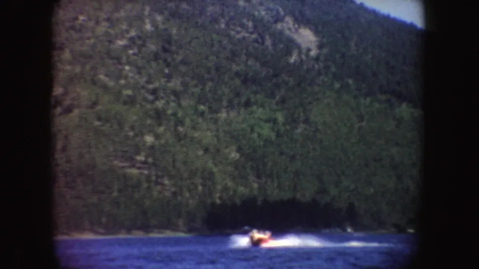 A small speedboat moves across a South Dakota lake in 1954 home-movie footage, with wooded hills behind it.