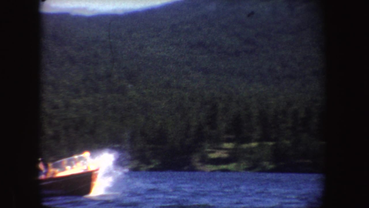 The speedboat appears closer at frame edge, cutting through blue water beneath a hazy forested hillside.