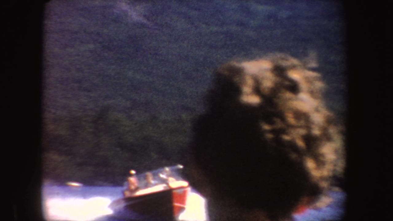 A speedboat passes on a blue lake while a blurred foreground head partly blocks the home-movie view.