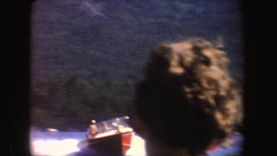 A speedboat passes on a blue lake while a blurred foreground head partly blocks the home-movie view.