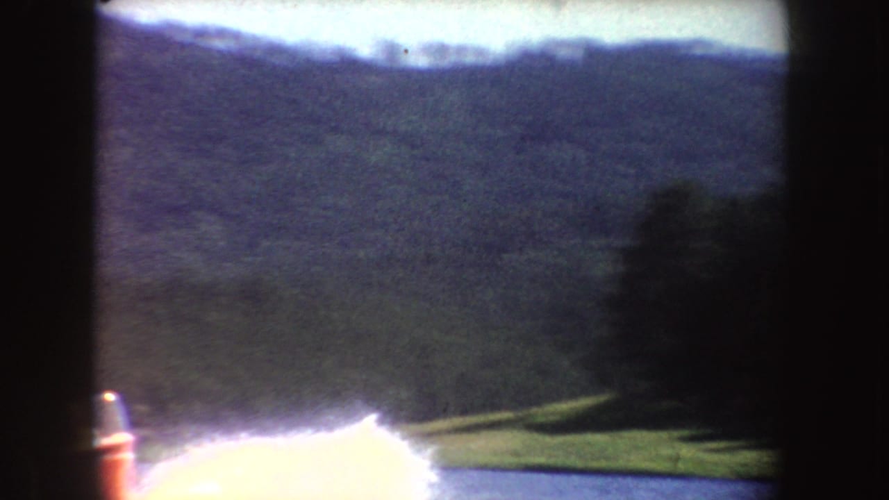 Boat wake at the edge of a lake with wooded hills in the background.