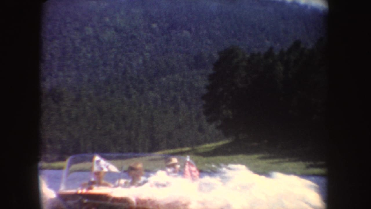 Passengers ride in a red motorboat on a South Dakota lake, with white wake, small flags, trees, and low hills visible behind them.