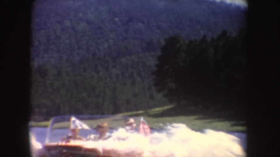 Passengers ride in a red motorboat on a South Dakota lake, with white wake, small flags, trees, and low hills visible behind them.