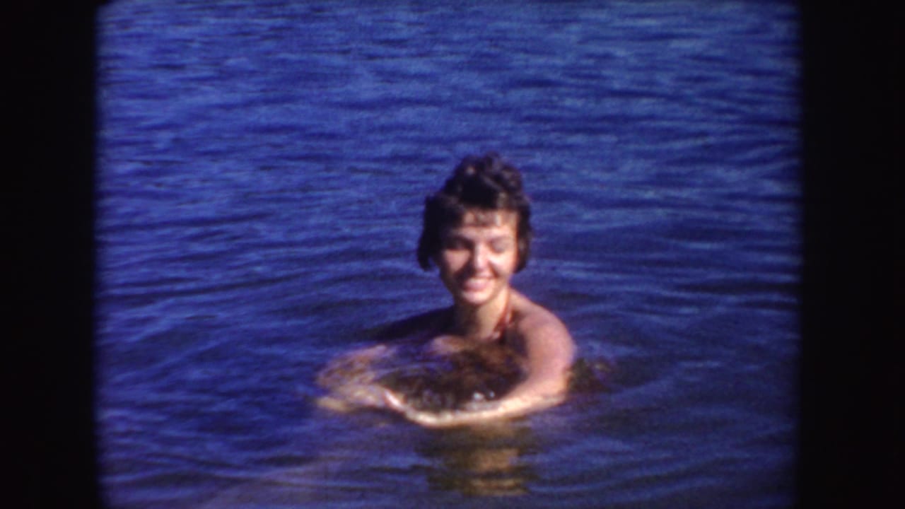 A smiling woman faces the camera while swimming in a South Dakota lake, shown in saturated 1954 8mm color.